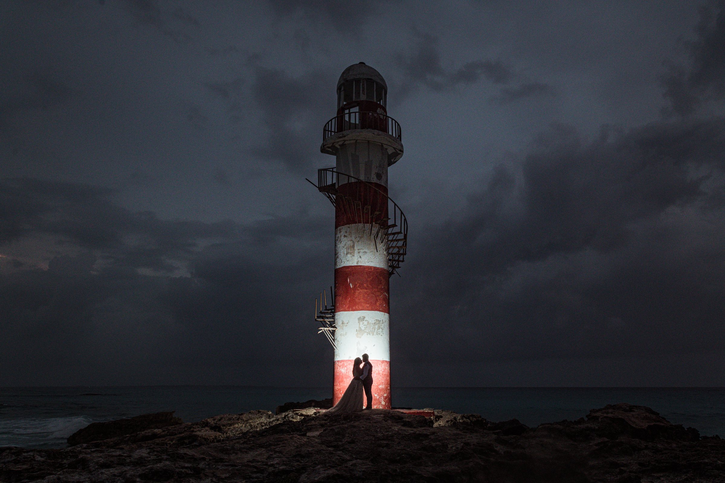 Los novios abrazados al pie de un faro iluminado, de noche