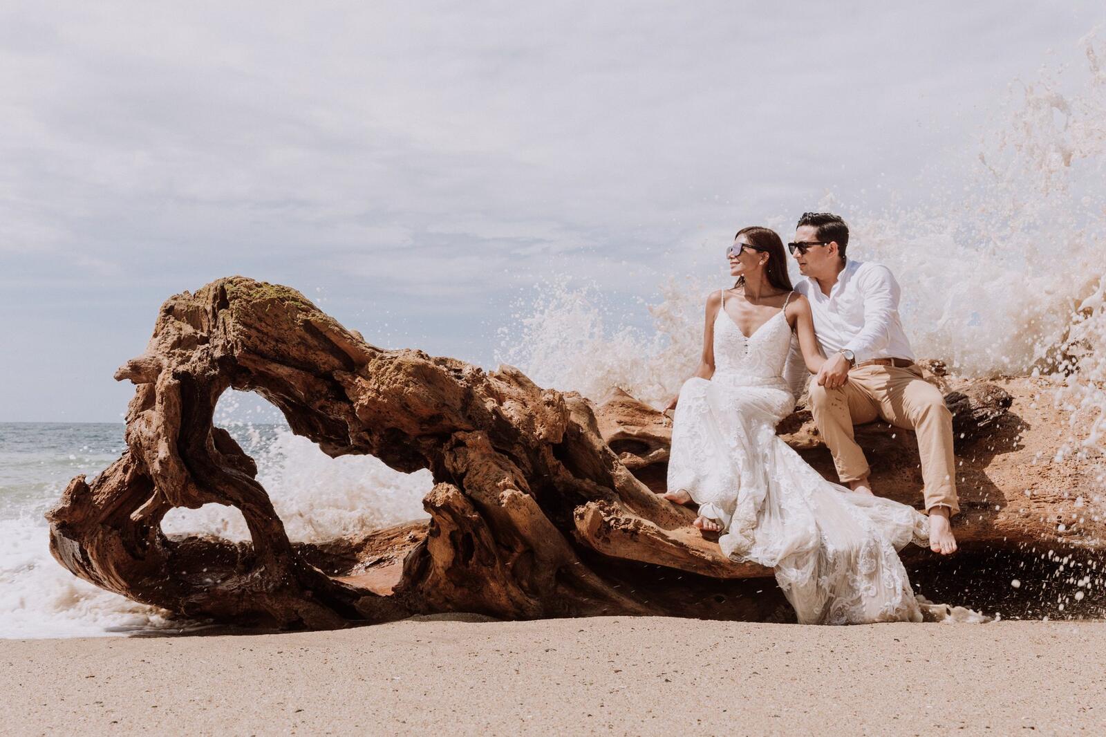 Los novios sentados sobre un tronco de madera a la orilla del mar