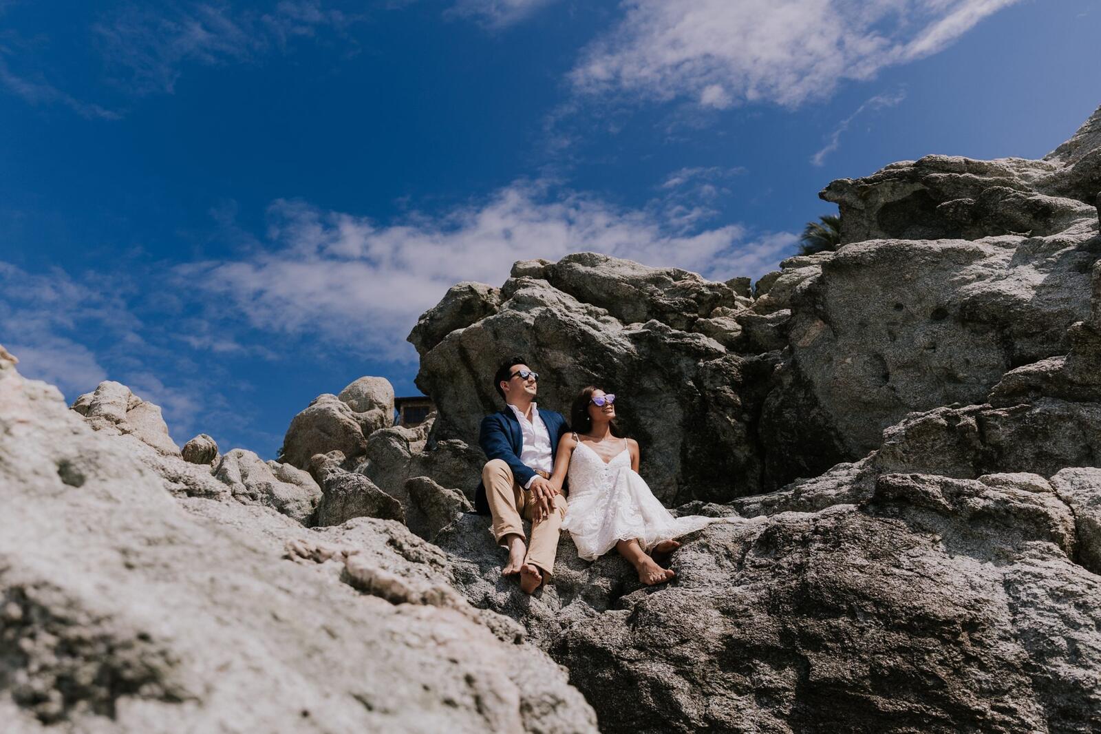 Los novios sentados en lo alto de unas rocas bajo el cielo azul