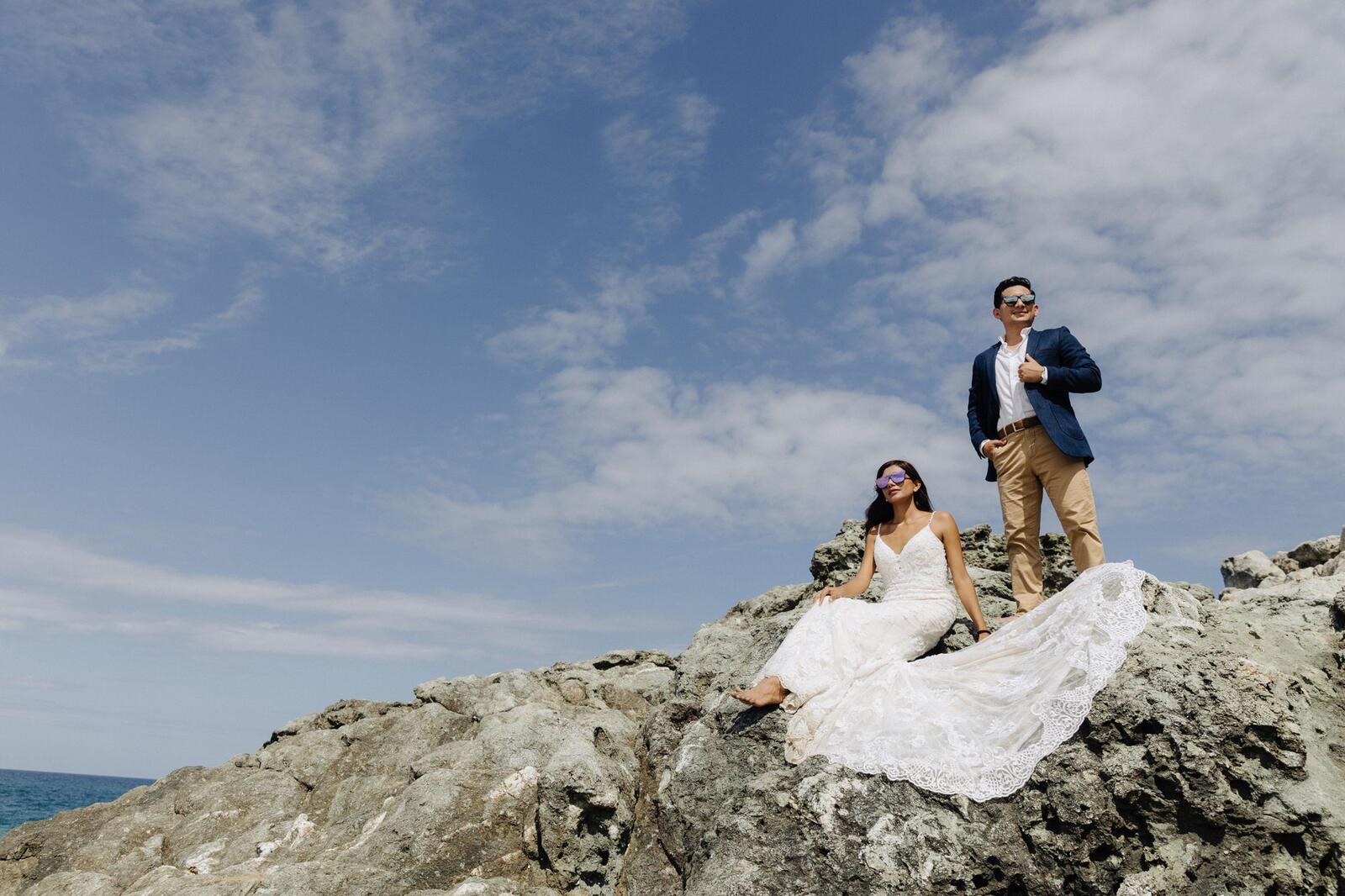 La novia sentada en la roca y el novio de pie, con el mar al fondo