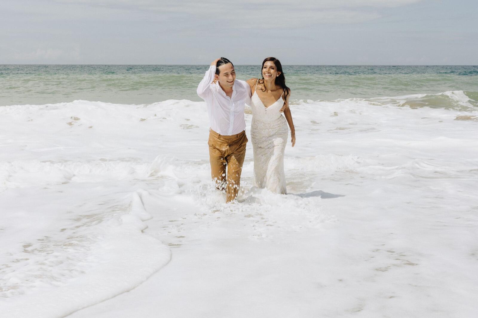 Los novios caminan riendo entre la espuma de las olas