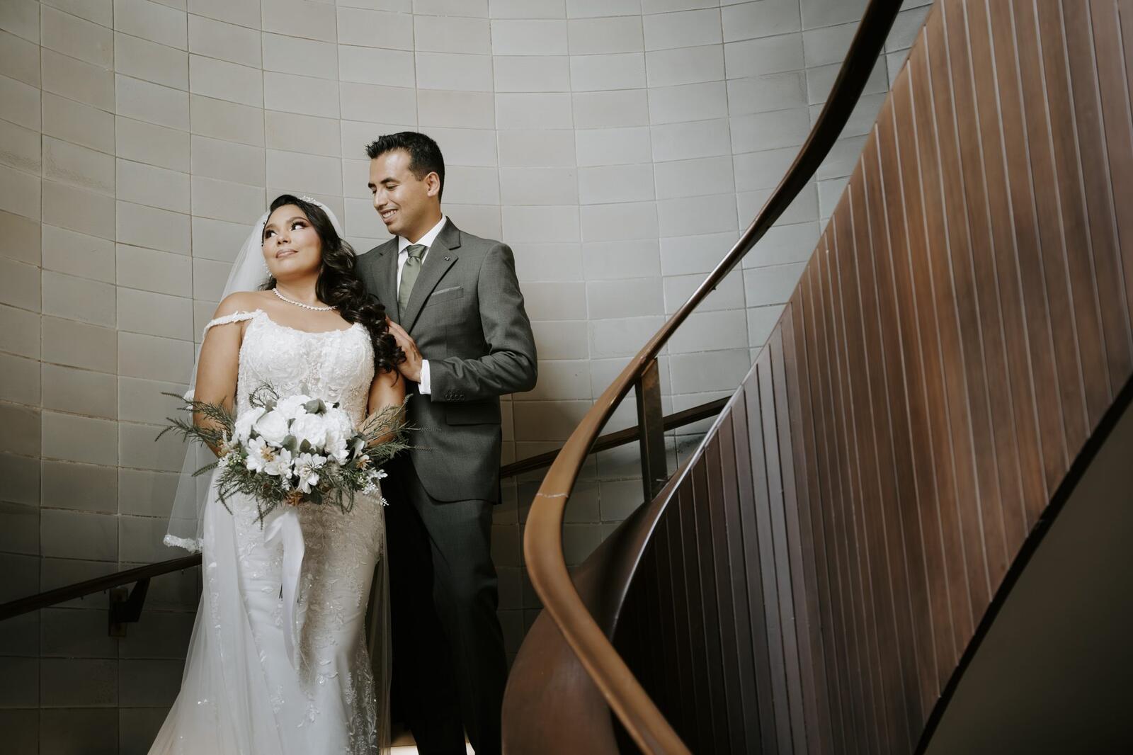 La novia con su ramo y el novio de traje gris junto a la escalera