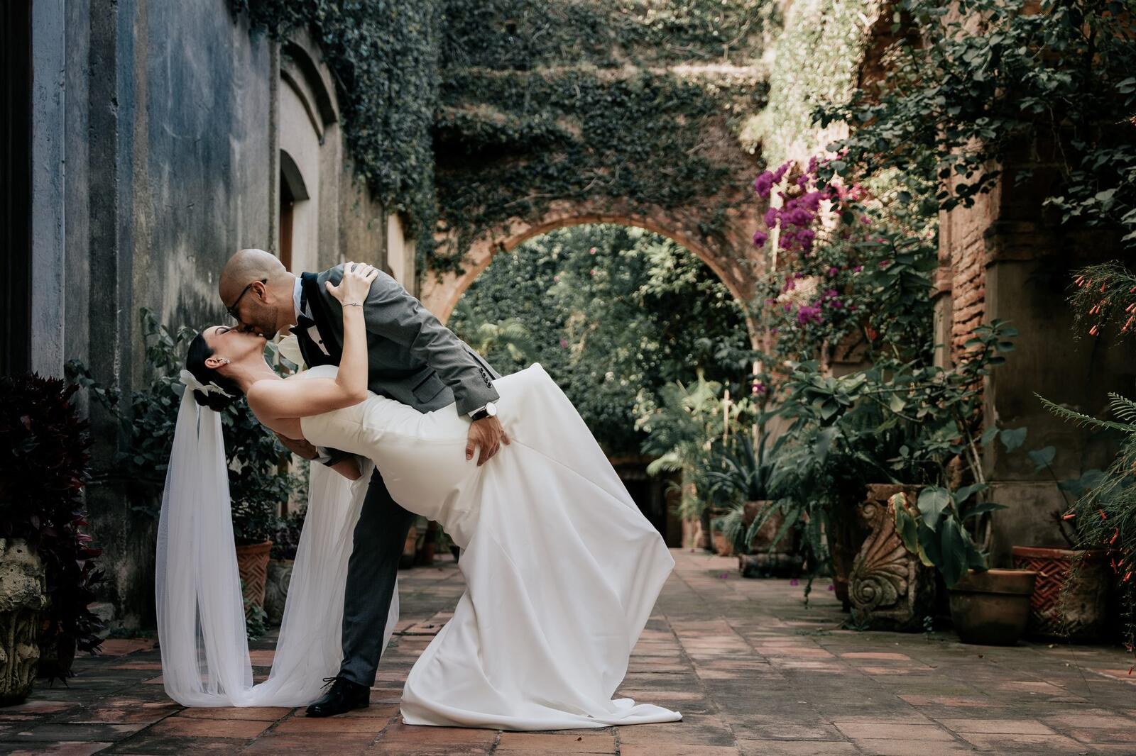 El novio inclina a la novia para besarla en un patio con arcos y bugambilias