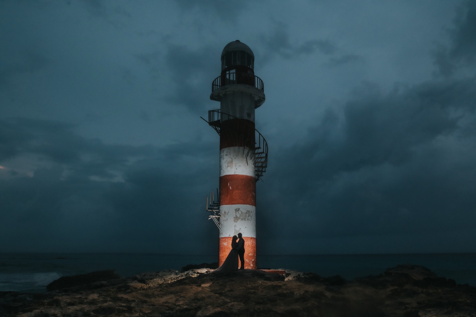 Los novios abrazados al pie de un faro rojo y blanco, al anochecer